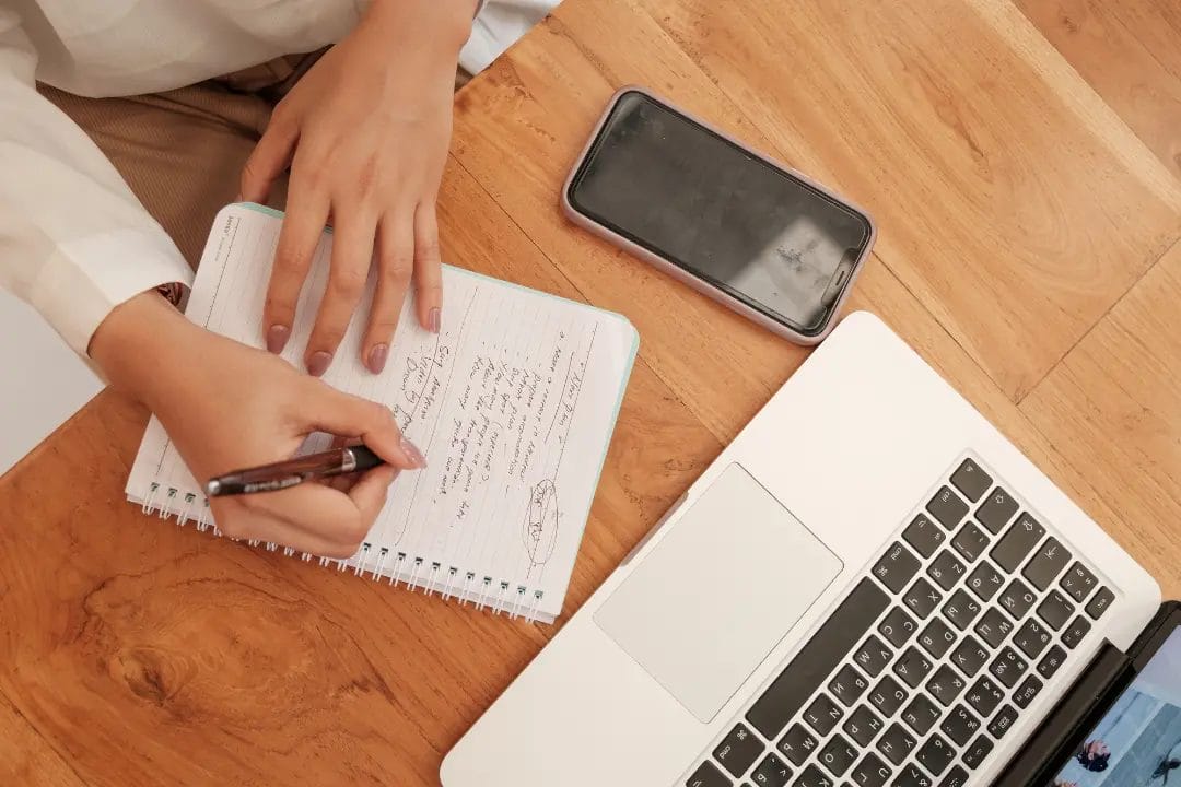 Person writing notes beside laptop and phone.